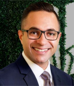 Professional portrait of a smiling man in a suit and glasses with green foliage background.