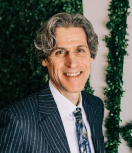 Professional portrait of Mark E. Noble, CPA in a suit with greenery background.